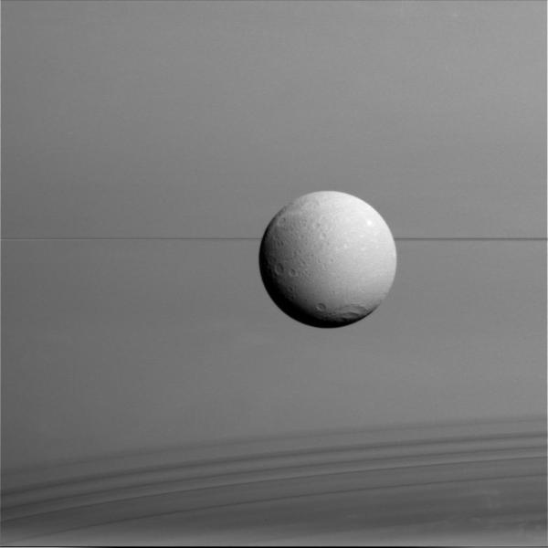 Dione imaged from a distance of 45,000 miles (73,000 km) on Aug. 17, 2015. Saturn fills the background, its edge-on rings cutting a thin dark line across the center with their shadows falling across the planet's clouds at bottom.